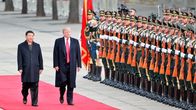 Presidentin Xi ja Trump kävelevät vakavina mustissa puvuissa ja päällystakeissa punaisella matolla kunniakaartin ohi. Xin takki on napitettu, Trumpin päällystakki on auki ja kirkkaanpunainen kravatti näkyy koko pituudeltaan.