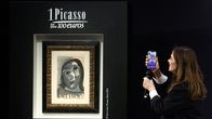 Picasson maalaus Tete de femme seinällä. Juontaja puhuu sen edessä videopuhelua.