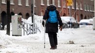 En person gående på en snöig gata med krycka och ryggsäck.
