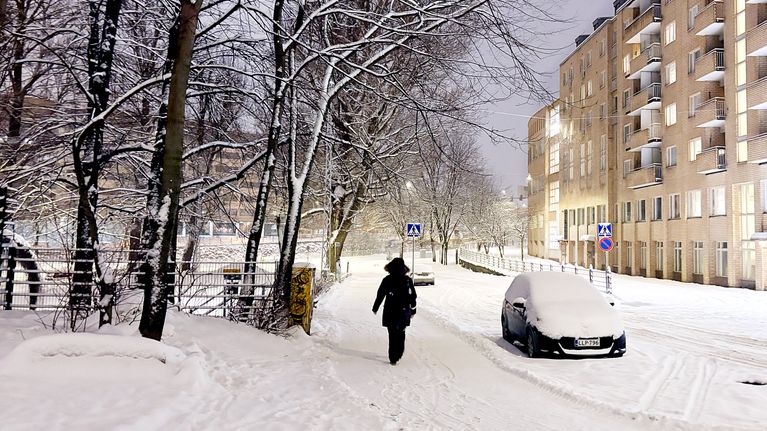 Henkilö kävelee talvivarusteissa lumisessa kaupunkiympäristössä. Auto on peittynyt lumeen.