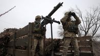Two Ukrainian soldiers in camouflage uniforms standing on the back of a truck, one looking through binoculars and the other behind a mounted machine gun.