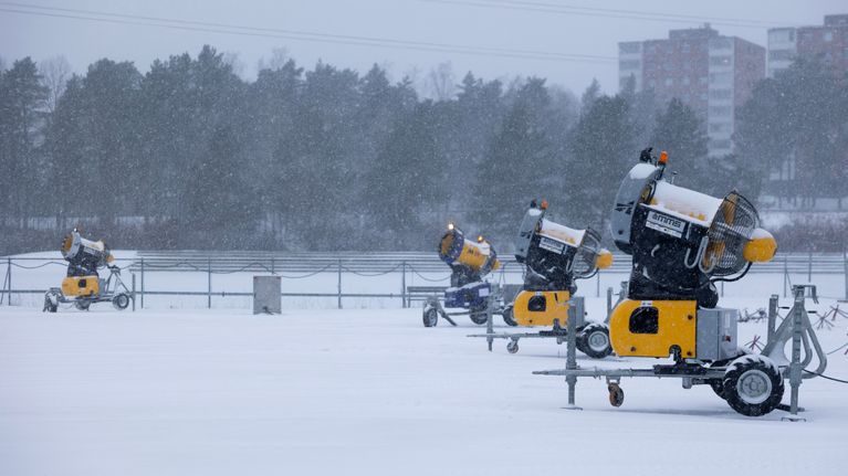 Neljä lumitykkiä, jotka eivät ole käytössä. Taivaalta pyryttää lunta. Taustalla on kerrostaloja. 