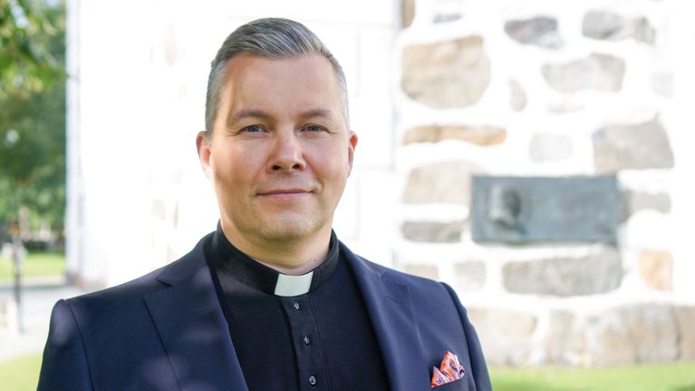 En person i prästkrage står utomhus framför en kyrkobyggnad med stenmurar och en plakett.