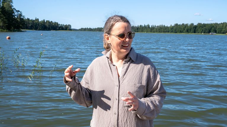 En person med solglasögon och randig skjorta står vid en sjö med skog i bakgrunden under klar himmel.