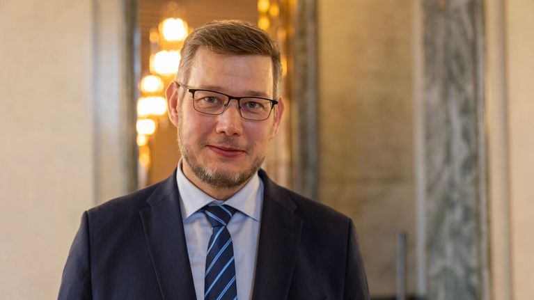 Silmälasipäinen nuorimies katsoo suoraan kameraan isossa aulassa.