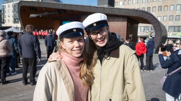Kaksi pitkähiuksista ihmistä hymyilee ja kaulailee torilla ylioppilaslakit päässään.