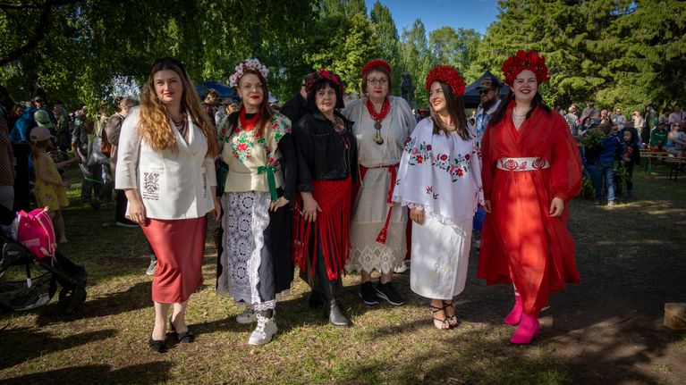 Six women stand in arm in arm in a park, wearing colourful traditional dresses and floral wreaths.