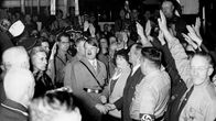 Adolf Hitler kanttajiensa keskellä kuvattuna. Takana on hymyileviä ja hailaavia natsipukuisia miehiä. 
