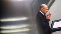Kuvassa näkyy Saksan liittokansleri Olaf Scholz, joka pitää puhetta, ja hänellä on edessään paperiarkkeja seisontapulpettia vasten. Taustalla on kirkasta valoa.