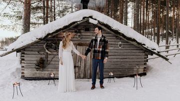 Valkoiseen pukeutunut morsian ja ruutupaitaan pukeutunut sulhanen katsovat toisiaan vanhan hirsiladon edessä.