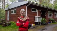 Seniori nainen seisoo ulkona punaisen, yksikerroksisen talon edessä sateisena kesäpäivänä.