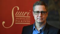 Silmälasipäinen mies seisoo Suuri Journalistipalkinto -tekstin edessä puku päällä ja hymyillen.