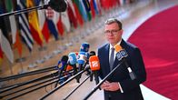 A man with brown hair and glasses and a blue suit stands before a cluster of microphones with a row of EU flags behind him.
