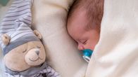 Infant boy sleeping under a white blanket next to a teddy bear.