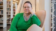 En kvinna sitter avslappnat i en fåtölj omgivet av böcker och dekorationer. Bakgrunden visar en trappa och bibliotekshyllor.