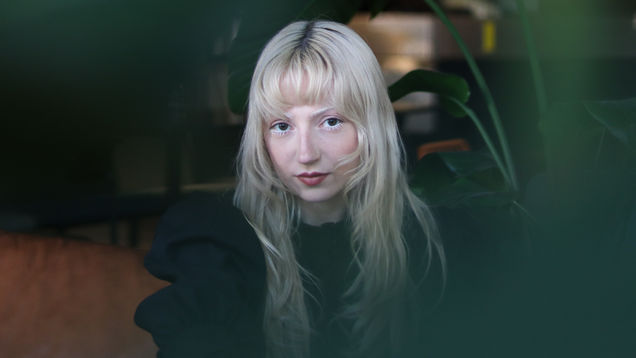 Muusikko Erika Sirola istuu sohvalla, etualalla isolehtinen kasvi.