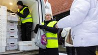 Marja Palkonen heijastinliivi päällään pakettiauton takana jakamassa ruoka-avustusta ja ojentamassa muovikassia jonottajalle.