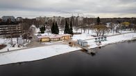 Talvinen maisema Tuiran uimarannalla, jossa on pieni rakennus ja saunarakennuksia. Taustalla näkyy kerrostaloja ja lumisia puita.