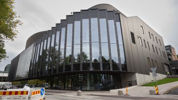 En stor, modern byggnad med svarta fasader och fönster i flera våningar. I förgrunden syns vägarbete med avspärrningar.