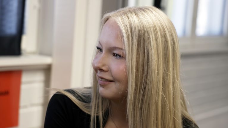 Vaaleatukkainen nuori nainen katsoo sivulle ja hymyilee koululuokassa. 