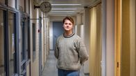 Herman Björk står i en korridor. Han bär en mönstrad tröja och jeans. Korridoren har ljusa väggar och gardiner till höger.