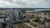Tampereen keskusta ja rakennuksia rautatieaseman läheisyydessä. Etualalla on rautatie ja junia, ja taustalla näkyvät modernit rakennukset sekä järvi ja vehreitä alueita. Taivas on pilvinen, mutta kirkas.