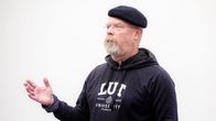 Man wearing eyeglasses, a black hoodie and beret standing in front of a blank whiteboard, gesturing with his right hand.