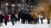 Ihmisiä kävelemässä talvisessa puistossa. Maassa on lunta ja puiston penkit ovat tyhjinä. Taustalla näkyy katuvaloja ja kaupungin rakennuksia.