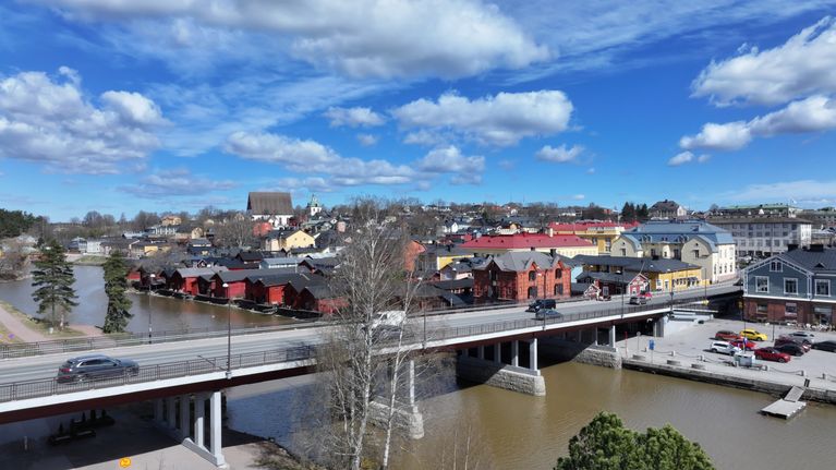 En bro leder över en å med bilar och byggnader på båda sidor under en blå himmel med moln.