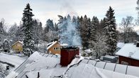 Snötäckt bostadsområde med röda och gula hus, rök stiger upp från skorstenen på ett tak.