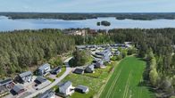 Ilmakuva pientaloalueesta, metsän ja pellon rajalta, taustalla järvi ja saaria kirkkaassa säässä.