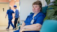 Nainen sinisissä työvaatteissa istuu nojatuolissa, taustalla kävelevät hoitaja ja kuntoutuja.