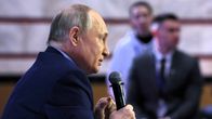 Tummapukuinen Vladimir Putin puhuu oikeassa kädessään pitämäänsä mikrofoniin. Taustalla näkyy häimeästi kuulijoita.