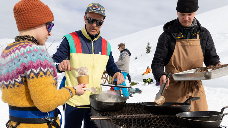 Three people wearing winter clothing stand around an outdoor grill on a sunny winter day, with others in the background.