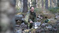 Henkilö on metsässä ja käyttää retkikeitintä. Hän hymyilee aurinkoisesti kameralle.