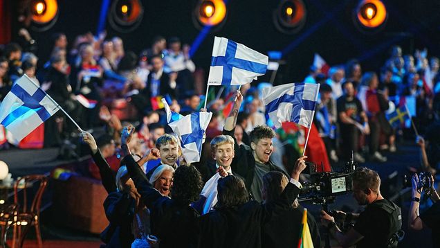 Ryhmä ihmisiä heiluttaa Suomen lippuja, taustalla näkyy yleisöä.