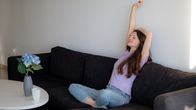 En kvinna i lila t-tröja sitter på en mörkgrå soffa och stretchar. Hon har ögonen stängda.