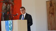 En man i kostym och slips i en talarstol, i bakgrunden hänger tavlor.