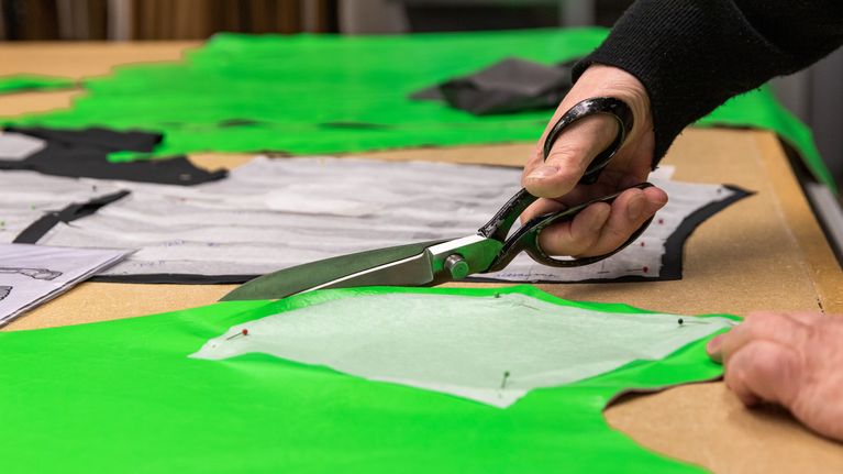 Kädessä olevat sakset leikkaavat vaaleanvihreää nahan palaa työpöydällä. Nahan päällä on vaaleasta paperista tehty mallipala.
