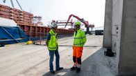 Two men in brightly coloured work clothes wearing helmets standing on a big concrete dock in front of a large cargo ship.