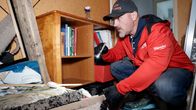 Mies punaisessa takissa ja lippalakissa tutkii taskulampulla huonetta, jossa on rojua ja likaa. Takana näkyy kirjahylly, jossa on kirjoja.