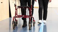 Mies kävelee rollaattorin kanssa liikuntasalissa nainen rinnallaan, kasvoja ei näy.