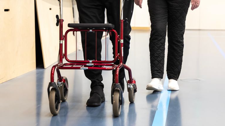 Mies kävelee rollaattorin kanssa liikuntasalissa nainen rinnallaan, kasvoja ei näy.