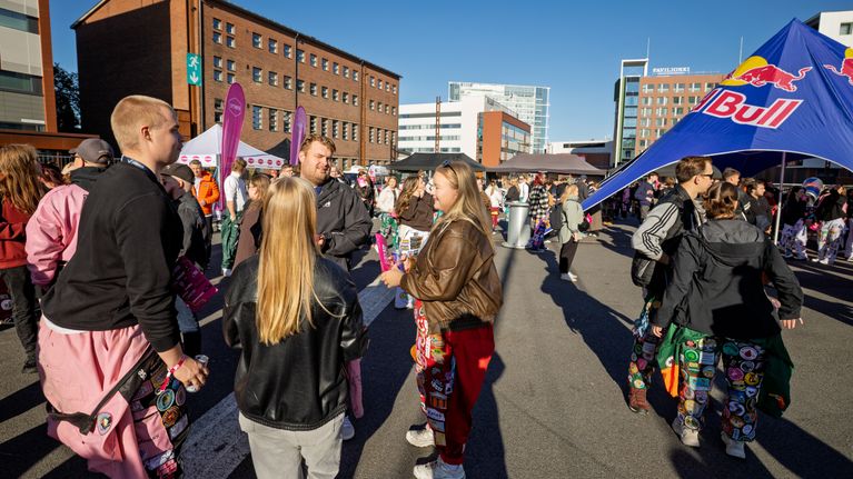 Ulkotilassa joukko haalareihin pukeutuneita nuoria seisoo keskustelemassa, taustalla Red Bull -teltta ja kerrostaloja.