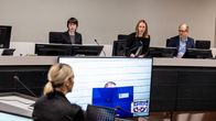 Oikeussalissa meneillään oleva oikeudenkäynti, jossa istuntosalissa on useita osallistujia ja suuri näyttö näyttää videoyhteyden välityksellä osallistujan.