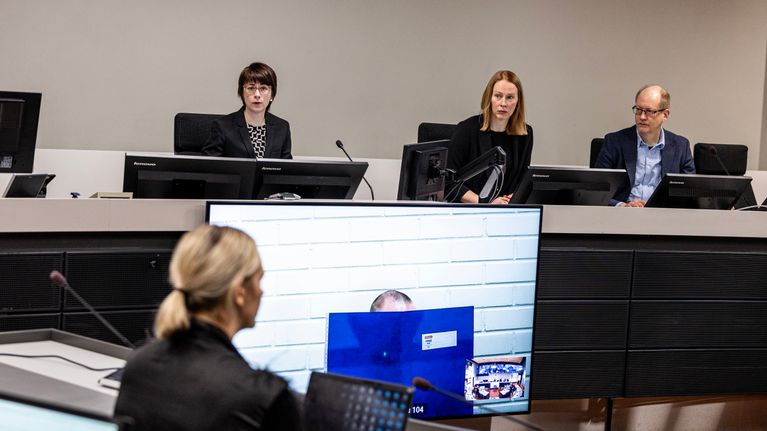Oikeussalissa meneillään oleva oikeudenkäynti, jossa istuntosalissa on useita osallistujia ja suuri näyttö näyttää videoyhteyden välityksellä osallistujan.