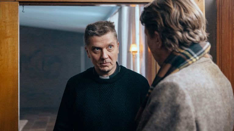 Pappi katsoo selin kameraan olevaa miestä sisätiloissa elokuvassa Pikku Siperia.