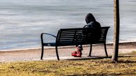 En person med luvtröja sitter på en bänk vid en strand. 