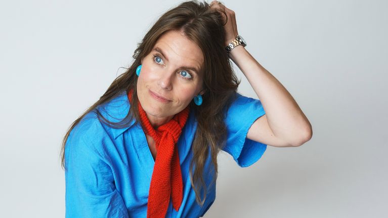 En person med blå t-shirt, röd scarf och jeans sitter på golvet mot vit bakgrund och håller en hand i håret.