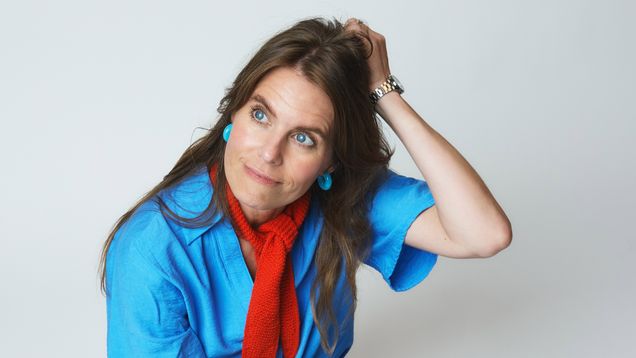 En person med blå t-shirt, röd scarf och jeans sitter på golvet mot vit bakgrund och håller en hand i håret.